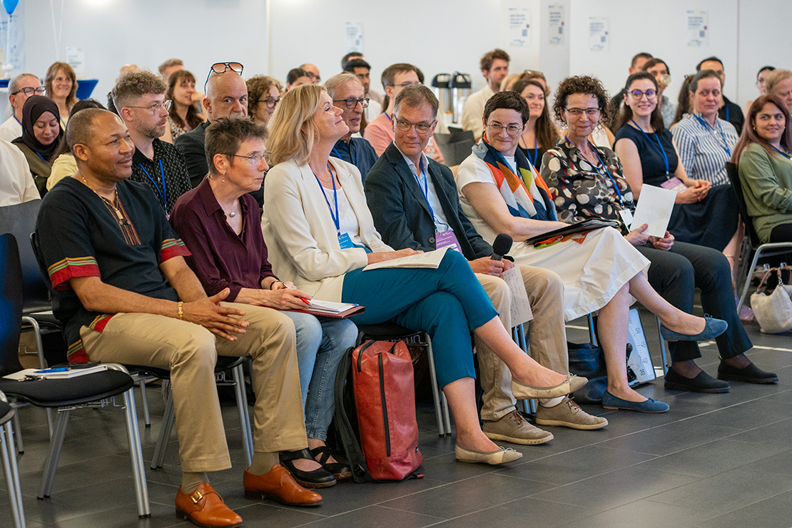 Konferenzpublikum sitzt aufmerksam in Stuhlreihen und hört einem Vortrag zu, mehrere Personen tragen Namensschilder, im Vordergrund macht sich eine Frau mit Notizblock Notizen