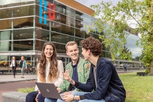 Studierende sitzen auf einer Mauer vor dem gläsernen Unigebäude an dessen Fassade das große blau-rote HU Logo zu sehen ist
