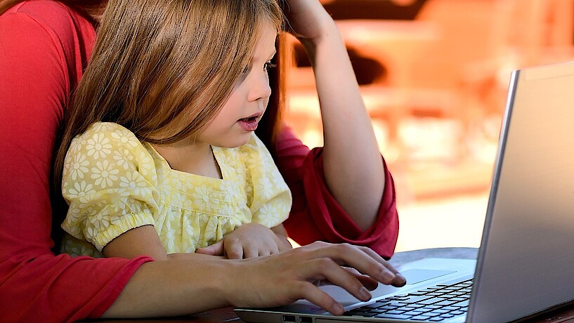 Ein kleines Mädchen sitzt auf dem Schoss der Mutter vor einem aufgeklappten Laptop an einem Schreibtisch