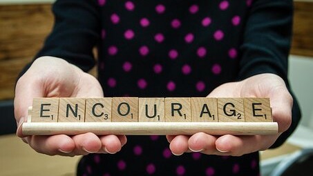 Das Foto zeigt ein Paar Hände, die Scrabble-Steine halten, die zusammen das Wort "Encourage", auf deutsch "Ermutigen" buchstabieren.