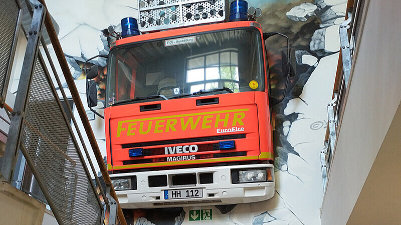 Treppenhausfoto mit einem Feuerwehrauto, dass aus der Wand zu fahren scheint.