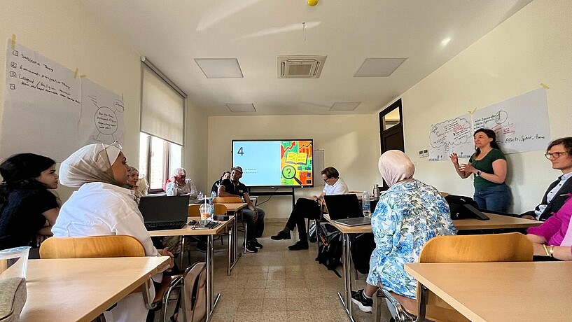 Ein Seminarzimmer mit Tischen und Stühlen, in denen eine Gruppe Menschen sich austauscht.