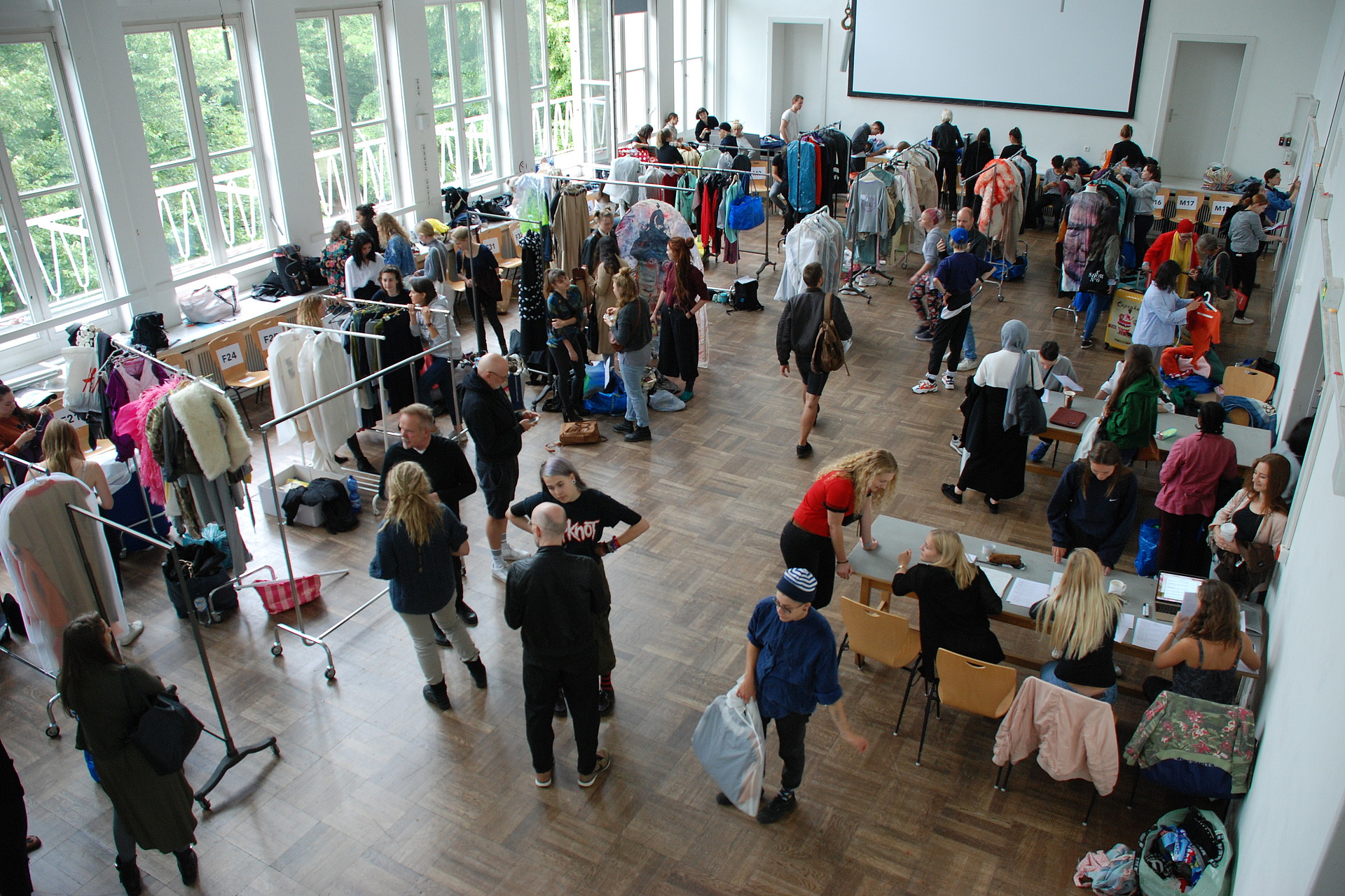 Bild von oben auf die Aula in der Armgartstraße, viele Menschen und Kleiderstangen mit Designstücken, die den Models angezogen werden.