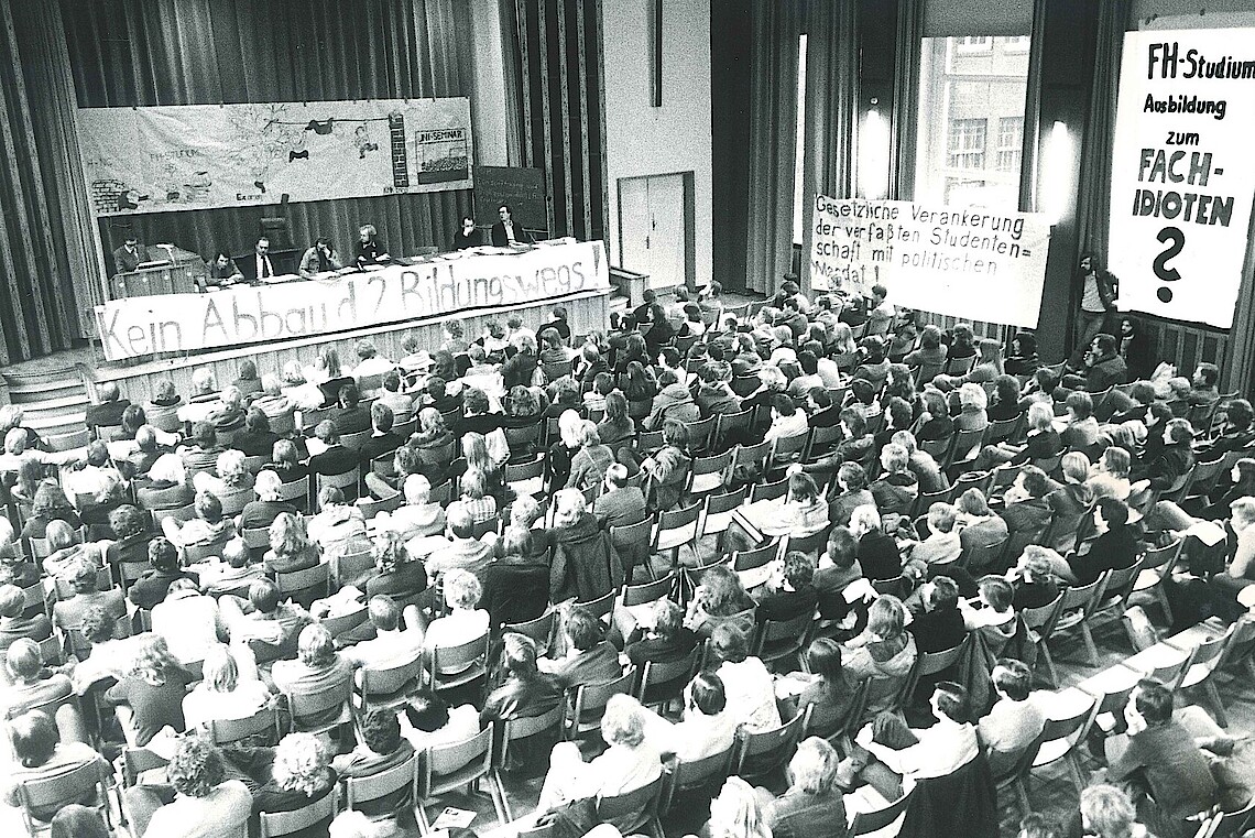 Blick von oben auf die voll besetzte Aula der FH Hamburg. Auf der Bühne steht ein langer Podiumstisch. Transparente sind auf der Bühne und an der Wand angebracht. 