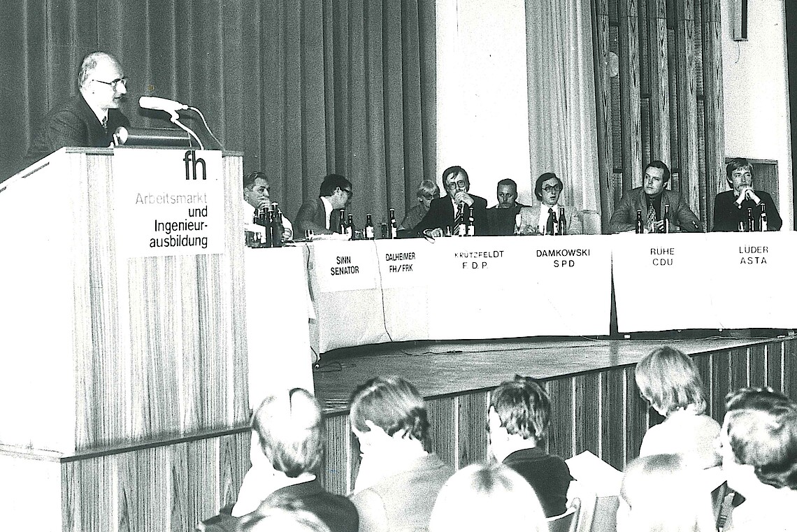 Schwarz-Weiß-Fotgrafie von einem Podium und Rednerpult auf einer Bühne. Am Rednerpult spricht ein Mann in das Mikrofon.