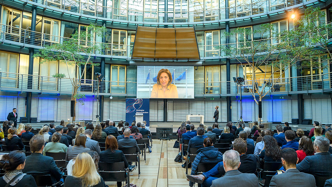 Das Bild zeigt das Plenum bei der HFD-Strategiekonferenz 2019 in Berlin. Das Publikum sitzt in einem Atrium. Auf der Bühne findet über eine Leinwand die Übertragung eines Grußwortes statt.