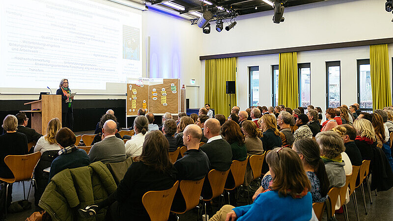 Präsentation auf der Bühne davor in mehreren Stuhlreihen Personen
