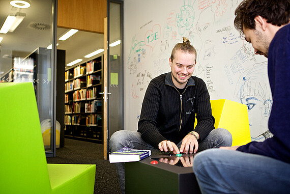 Zwei Studierende sitzen sich gegenüber auf bunten Sitzgelegenheiten und spielen ein Computerspiel, das zwischen ihnen steht. 
