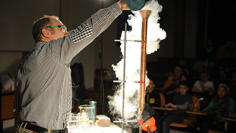 Prof. Dr. Marcus Schiefer gießt Flüssigkeit in ein Rohr und daraus entsteht Dampf. Die Kinder im Hintergrund finden es spannend.