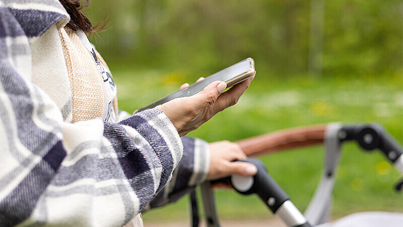 Person hält in der rechten Hand ein Smartphone und in der linken Hand den Griff eines Kinderwagens