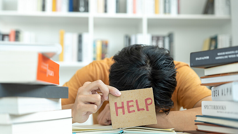 Eine Frau legt den Kopf zwischen auf den Schreibtisch und hält eine Pappschild mit der Aufschrift "HELP" hoch
