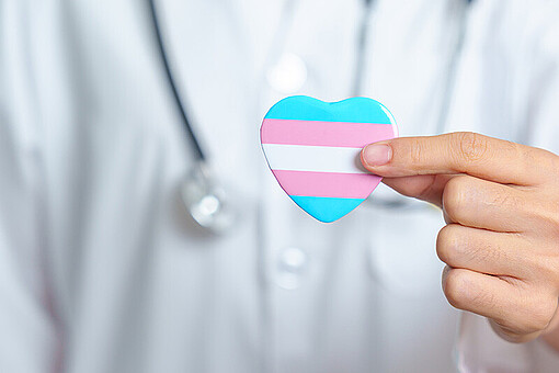 
                Herz mit Farben der trans* Flagge vor einer Person in einem medizinischen Kittel mit Stethoskop
            
