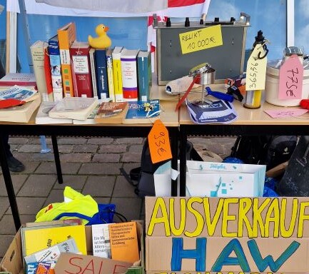 Das Foto zeigt den Ausschnitt eines Flohmarktstandes "Ausverkauf der HAW", wo verschiedene Artikel, die mit der Hochschule verbunden werden, zum Verkauf angeboten werden.