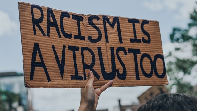 auf dem Foto hält eine Hand ein Papp-Schild hoch, auf dem in schwarzer Schrift steht "Racism is a Virus too" (deutsch: Rassismus ist auch ein Virus).