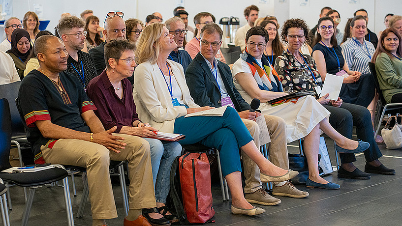 Fachtagung26-03 Konferenzpublikum sitzt aufmerksam in Stuhlreihen und hört einem Vortrag zu, mehrere Personen tragen Namensschilder, im Vordergrund macht sich eine Frau mit Notizblock Notizen