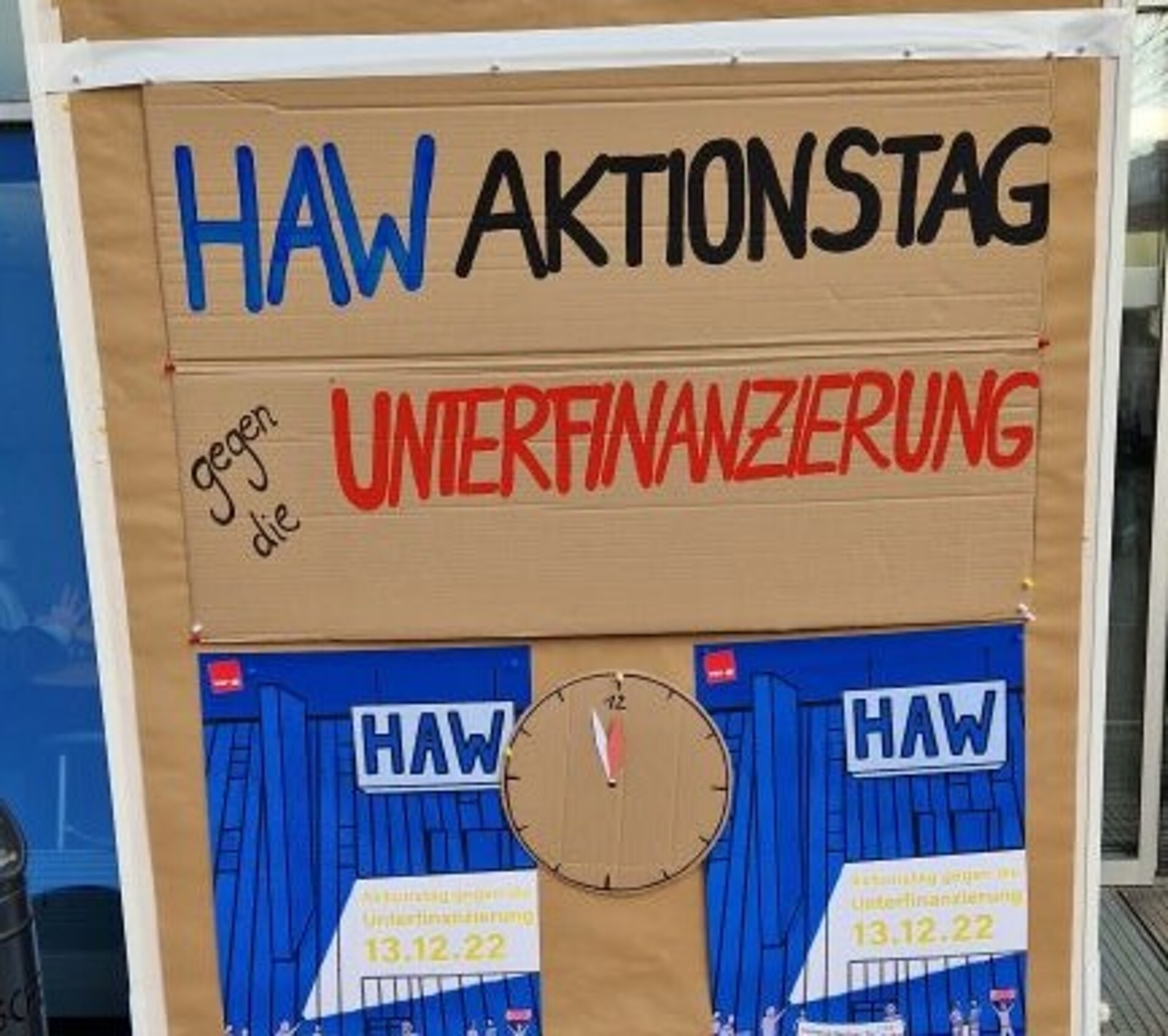 Das Foto zeigt eine Stellwand mit der Überschrift: HAW Aktionstag gegen die Unterfinanzierung, sowie eine Uhr, die 5 Minuten vor 12 Uhr anzeigt.