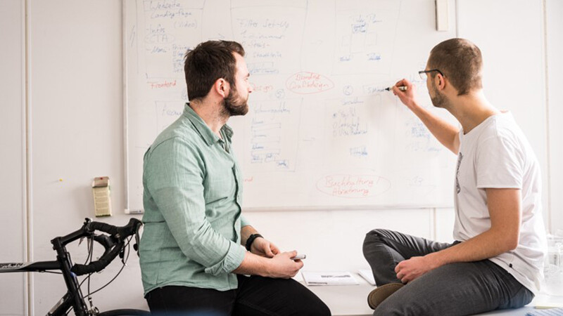 Zwei Männer sitzen sich auf Schreibtischen gegenüber, hinter ihnen ist ein Whiteboard, auf dem der Mann rechts im Bild etwas schreibt