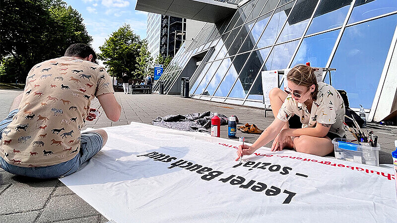 Zwei Studierende vor dem Gebäude des Berliner Tor 5, sitzend auf dem Steinboden, beschriften ein Demonstrations-Plakat