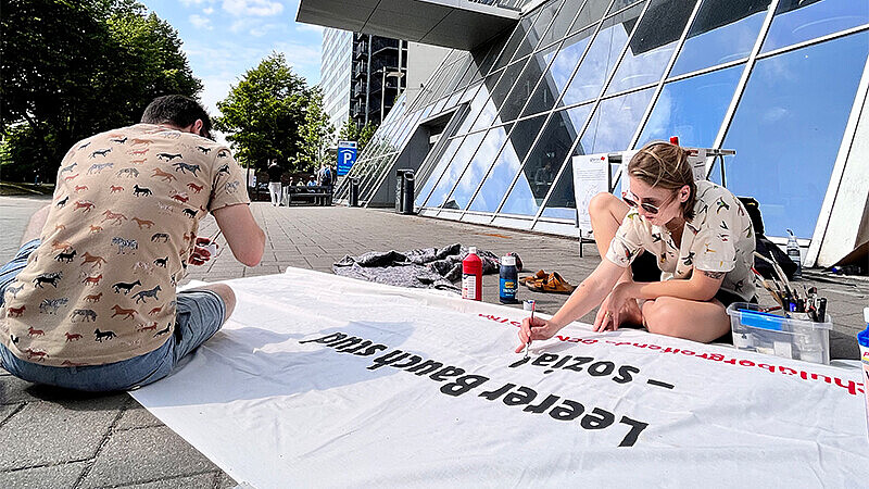 Zwei Studierende vor dem Gebäude des Berliner Tor 5, sitzend auf dem Steinboden, beschriften ein Demonstrations-Plakat