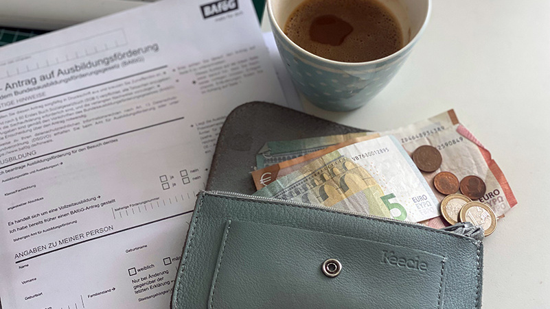 Ein offenes Portemonnaie mit Geldscheinen und Münzen. Im Hintergrund eine Tasse Kaffee und ein BAföG-Antrag