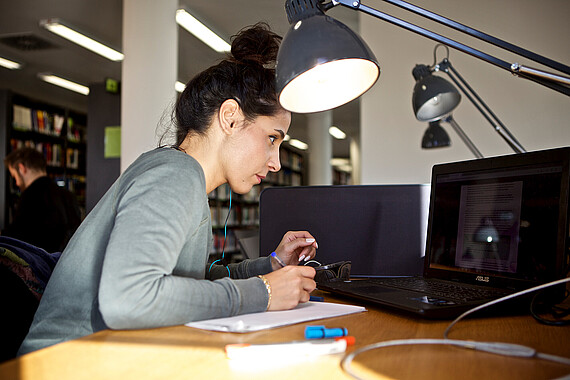 Eine Frau sitzt vor ihrem Laptop unter einer Lampe an einem Arbeitsplatz in der Bibliothek.