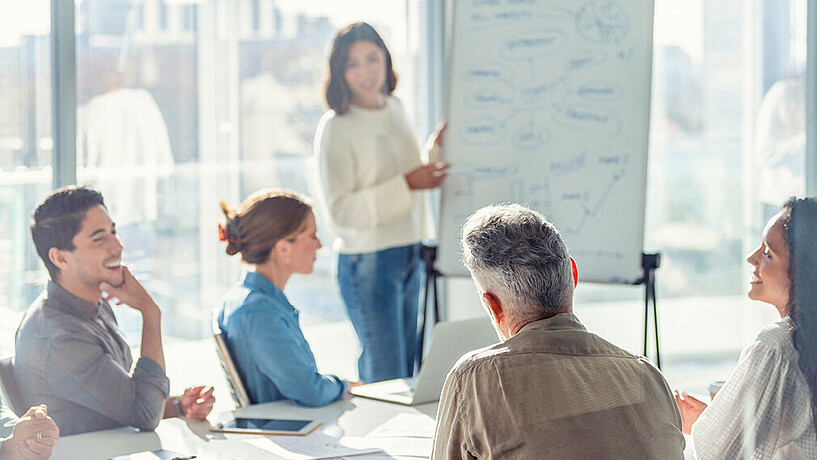 Personen im Seminarraum an einem Tisch, Person stehend neben Flipchart