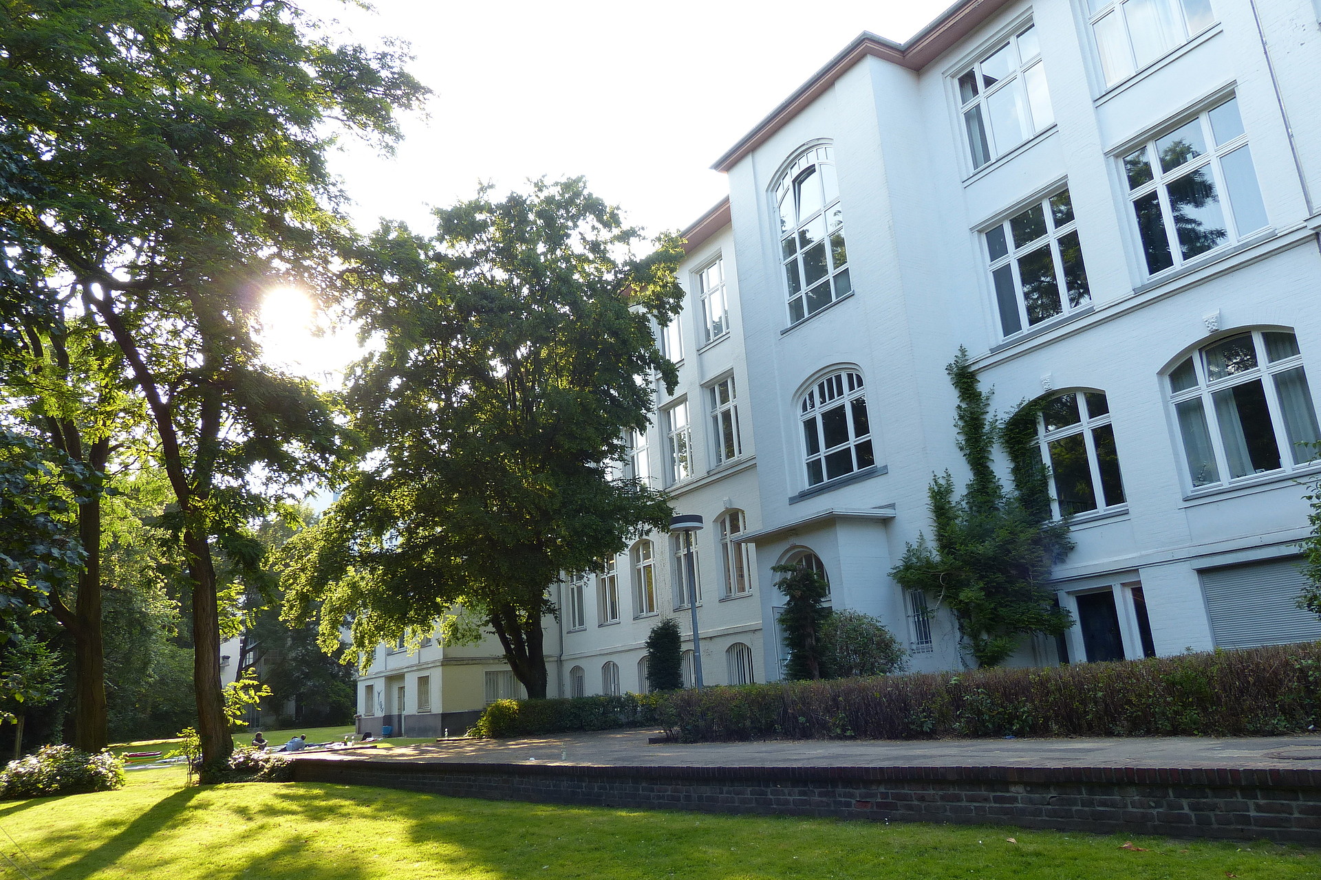 The sun shines through the green trees in a garden in front of the white building of the Armgartstraße Campus