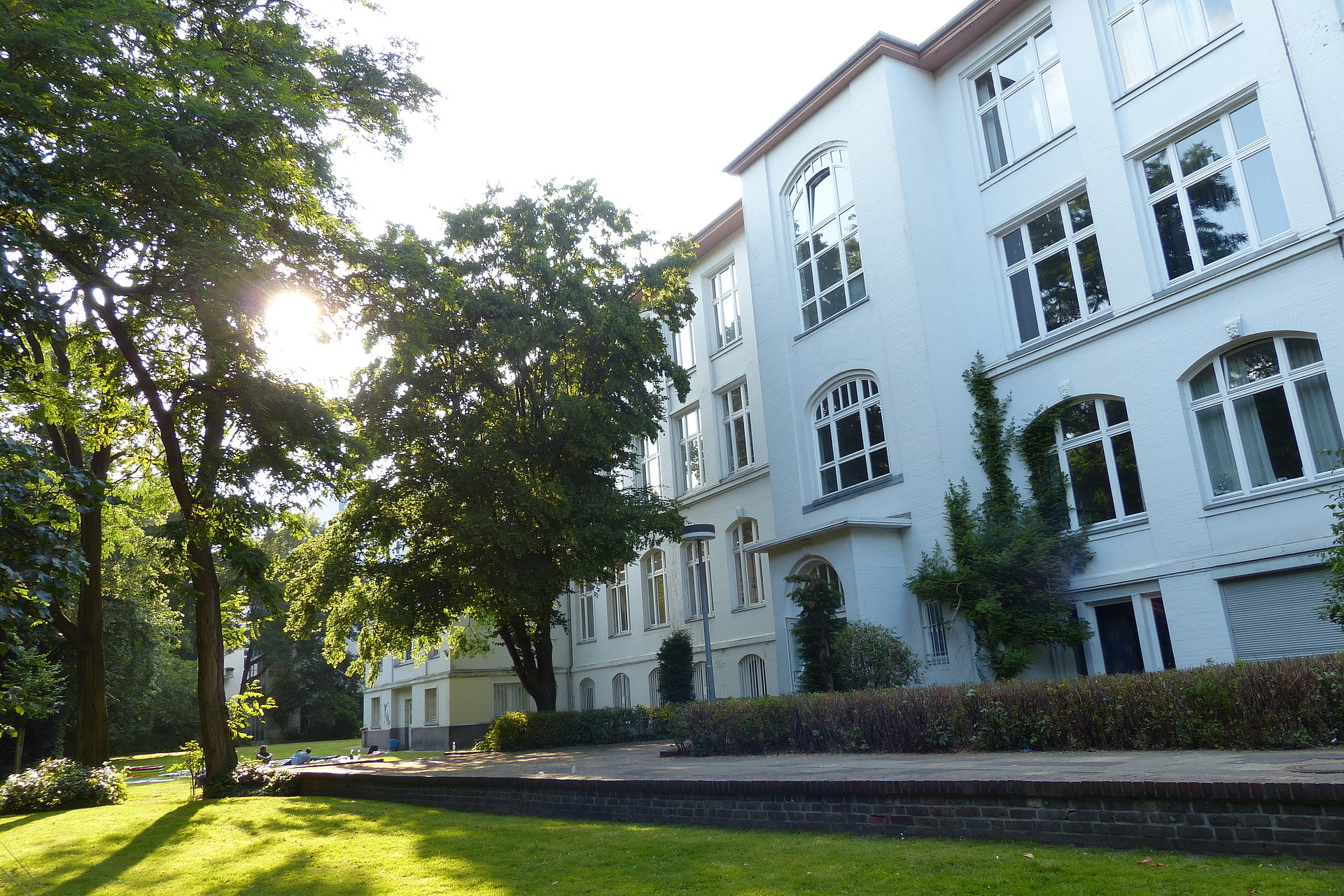 Die Sonne scheint durch die grünen Bäume in einem Garten vor dem weißen Gebäude in der Armgartstraße