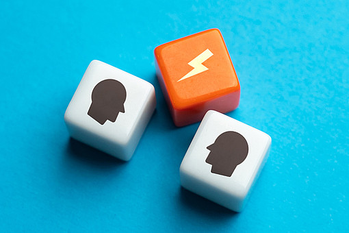 
                Zwei Gesichter auf weißen Bausteinen sind einander zugewandt, in der Mitte befindet sich ein orangener Baustein mit einem Gewitterzeichen.
            