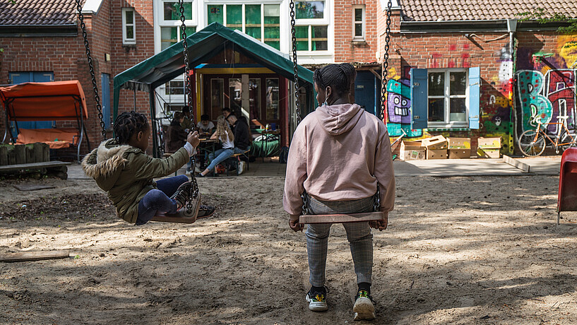 Zwei Mädchen schaukeln auf einem Spielplatz und mehrere Kinder und Jugendliche sitzen in einem kleinen Spielplatz-Häuschen. Im Hintergrund sieht man das Gebäude einer Kinder- und Jugendeinrichtung.