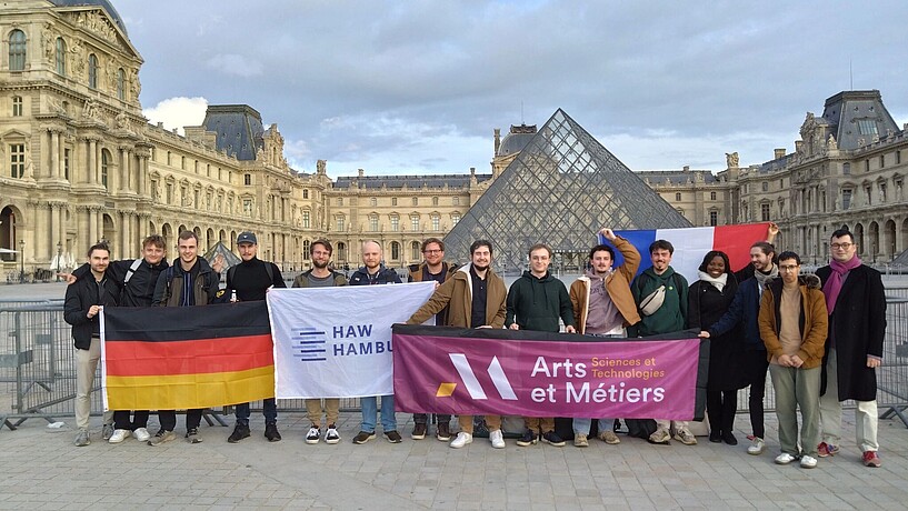 Teilnehmer:innen des Hackathons stehen vor dem Louvre in Paris und halten Flaggen der beiden Hochschulen, sowie die Deutsche und die französische.